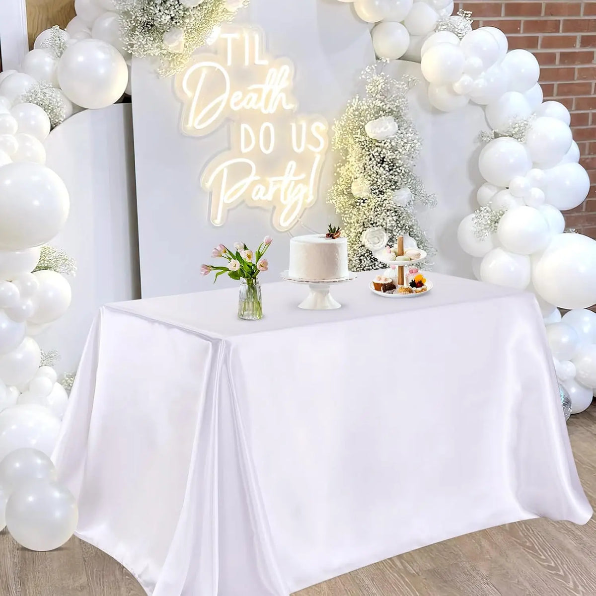 A white satin tablecloth is draped on a table, set for an elegant party with a cake and floral decorations.