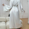 A woman in a white silk satin robe with lace trim is shown from the back, standing in a bright room.