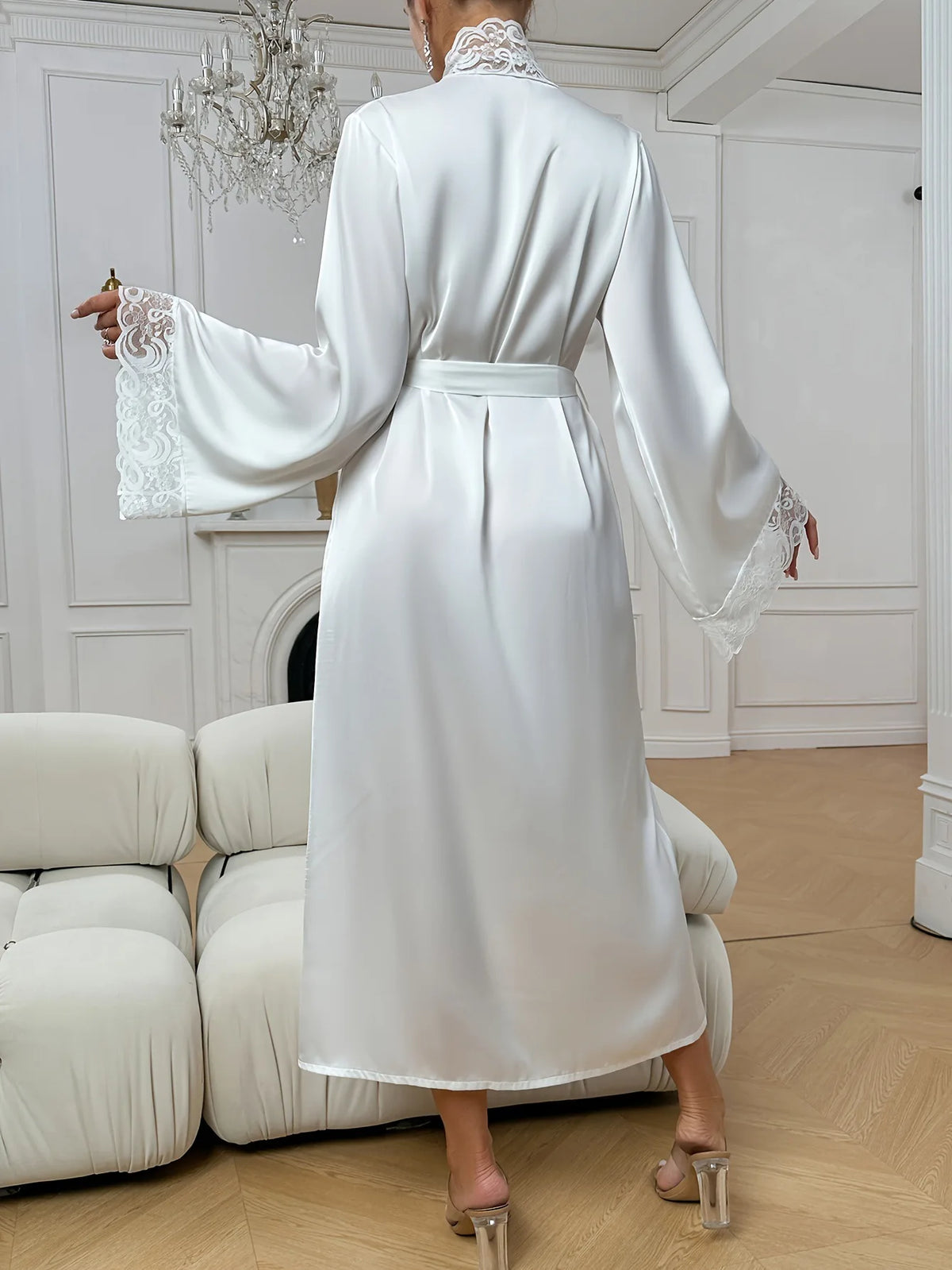 A woman in a white silk satin robe with lace trim is shown from the back, standing in a bright room.
