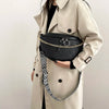 A woman is holding a black woven pattern waist bag with gold chain details against a light beige trench coat.