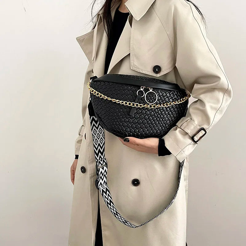 A woman is holding a black woven pattern waist bag with gold chain details against a light beige trench coat.