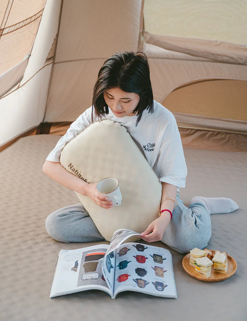 A person is reading a book while resting their head on a beige foam inflatable camping pillow in an outdoor tent setting.