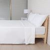 A white microfiber bed sheet set is neatly folded on a wooden bed frame next to a bedside lamp.
