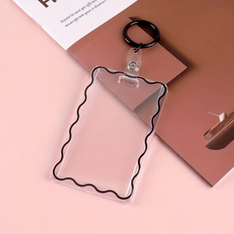 Porta tarjetas de acrílico transparente con borde decorativo unido a un llavero negro, sobre una superficie rosa.