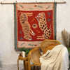 A woven blanket featuring a red background with wildlife motifs like a kangaroo and leopard, displayed on a white wall.