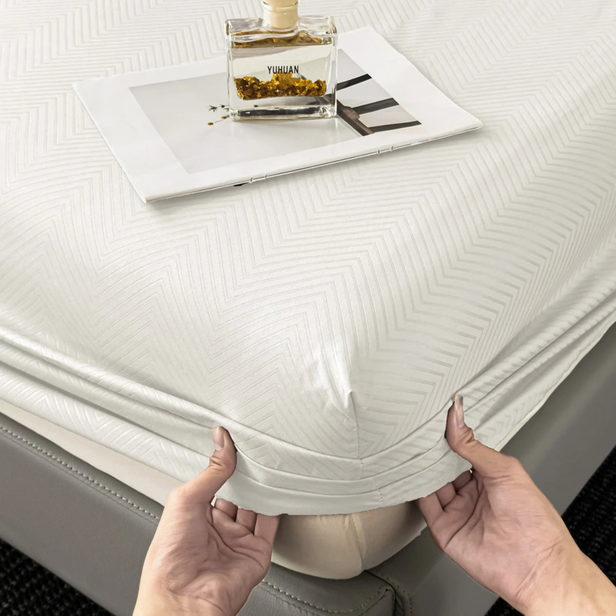 A white fitted sheet is displayed on a bed, with a glass bottle of perfume resting on the sheet.