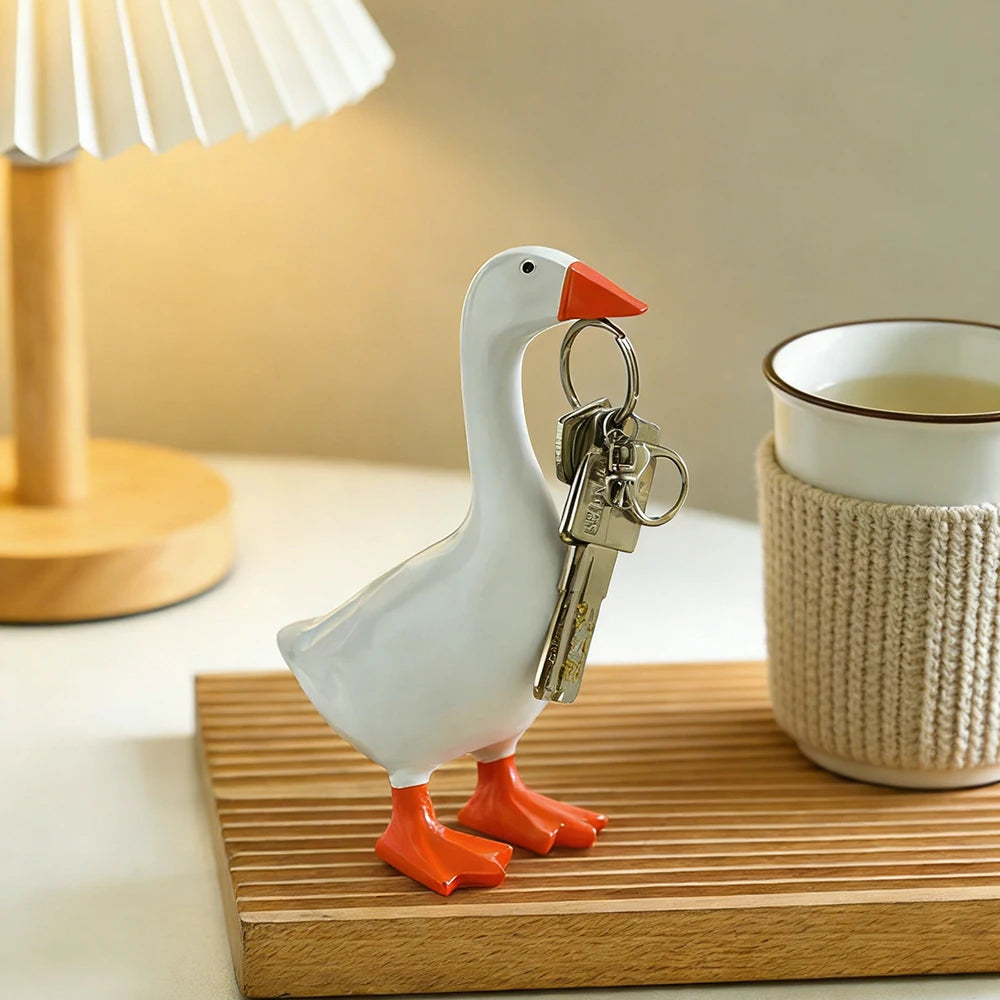 A white resin goose sculpture with orange beak and feet holding a set of keys on a wooden desk.