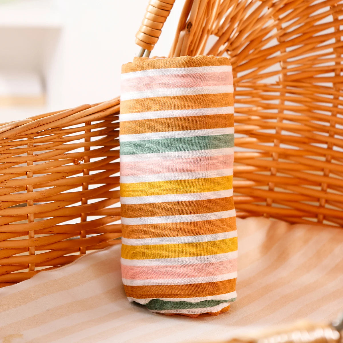 A striped orange and pastel muslin baby swaddle blanket is displayed next to a woven basket.