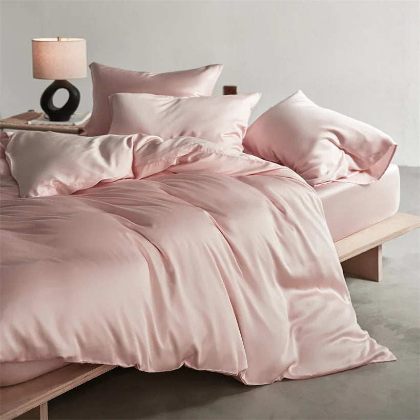 A pink luxury mulberry silk duvet cover set displayed on a bed with matching pillows and a bedside lamp.