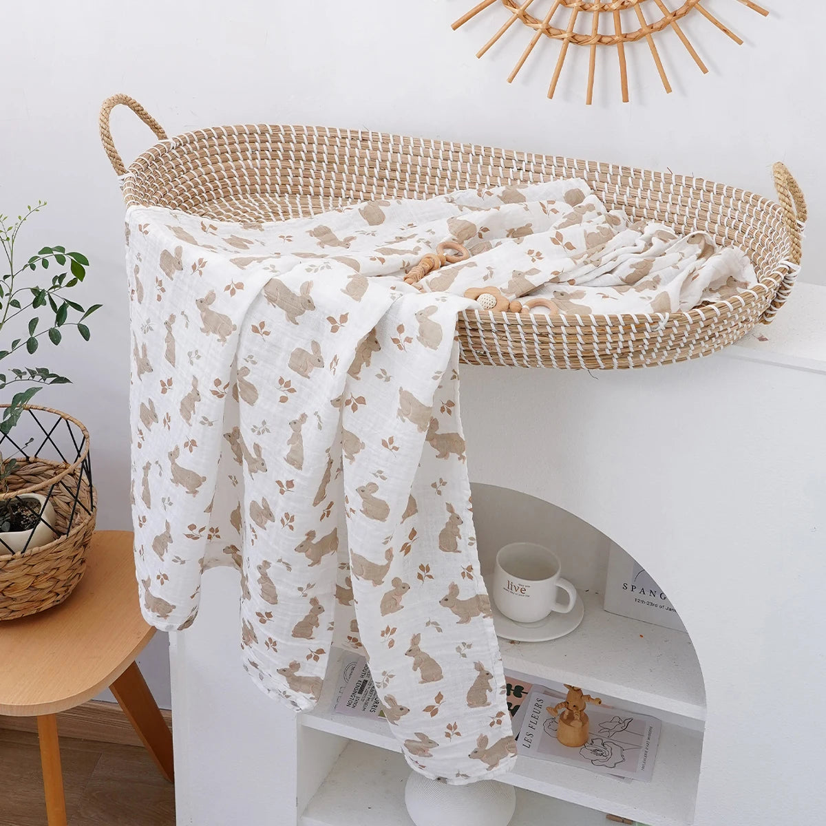 A cream and beige patterned muslin baby swaddle blanket draped over a woven basket on a white shelf.