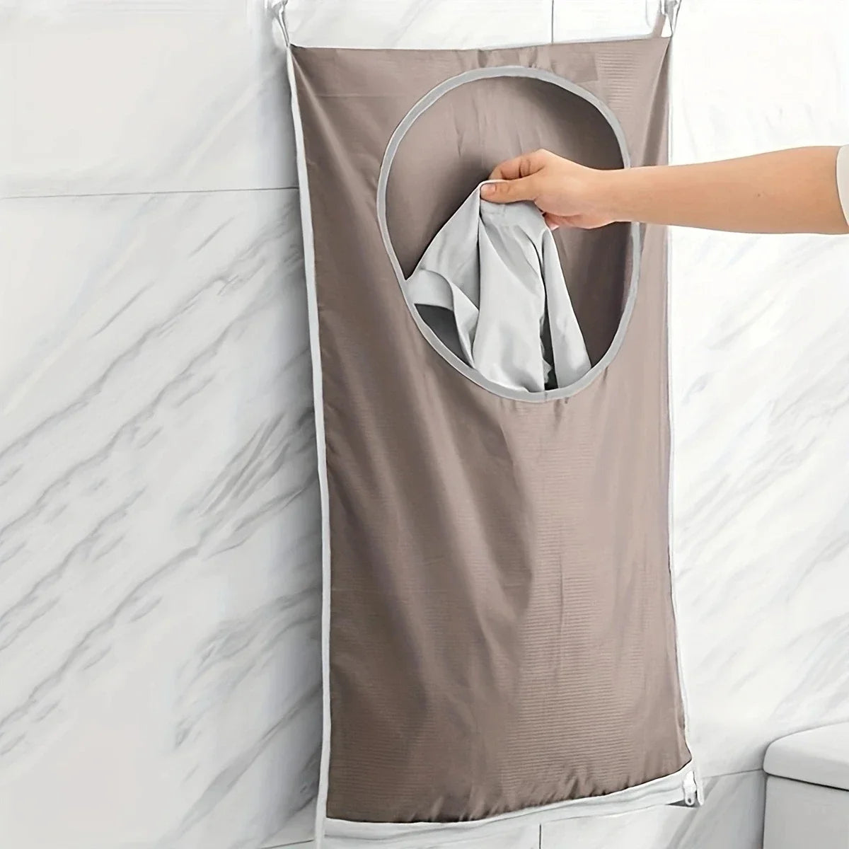 A hand is pulling out a folded gray towel from a fabric laundry storage bag hanging on a white marble tile wall.