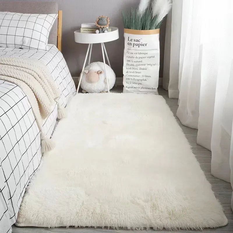 A fluffy white silk-touch area rug placed on a light wooden floor next to a bedside table.