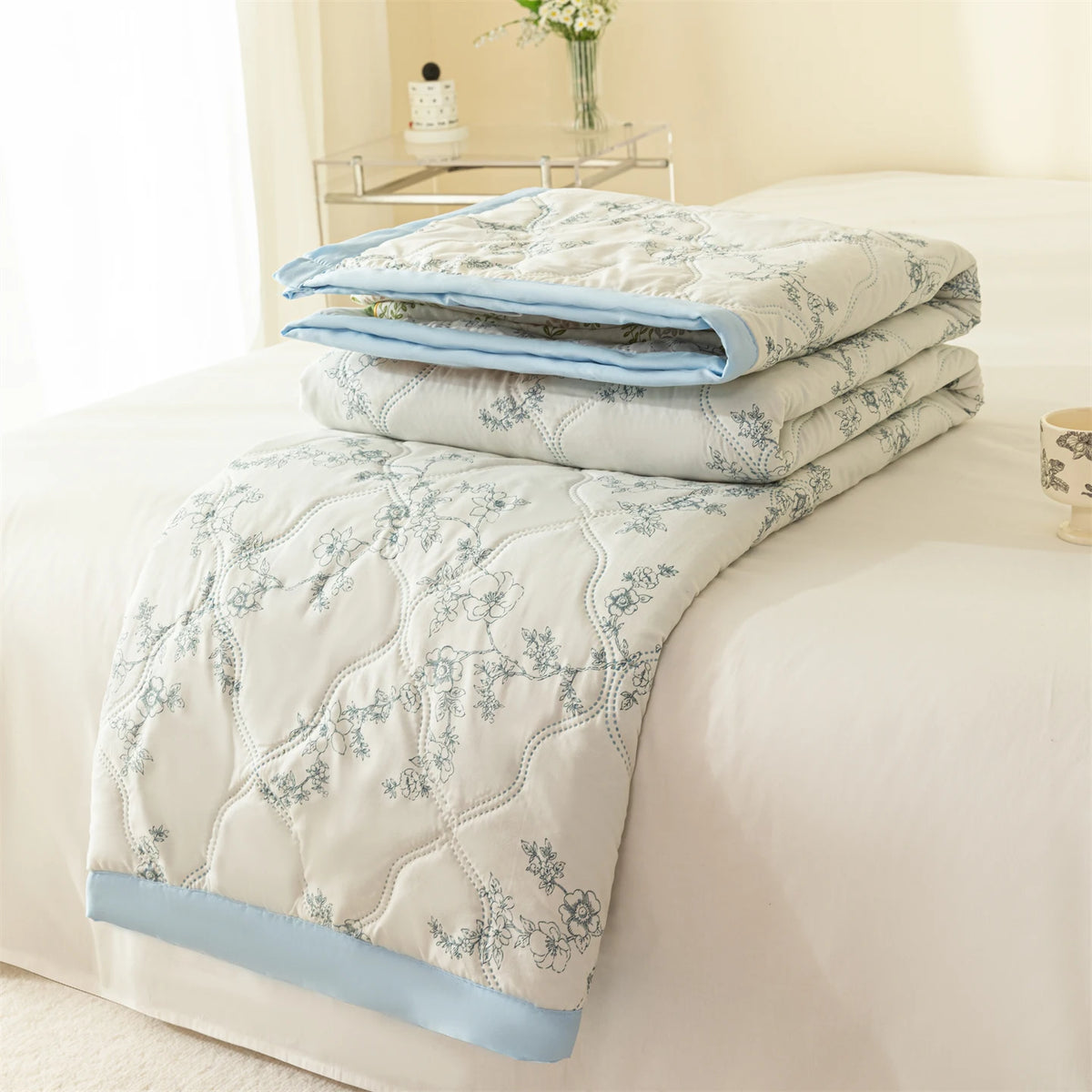 A light blue quilted blanket folded on a white bed with a pale floral pattern.