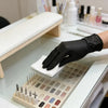 A hand wearing a black disposable nitrile glove is placing cosmetic applicators on a white tray.