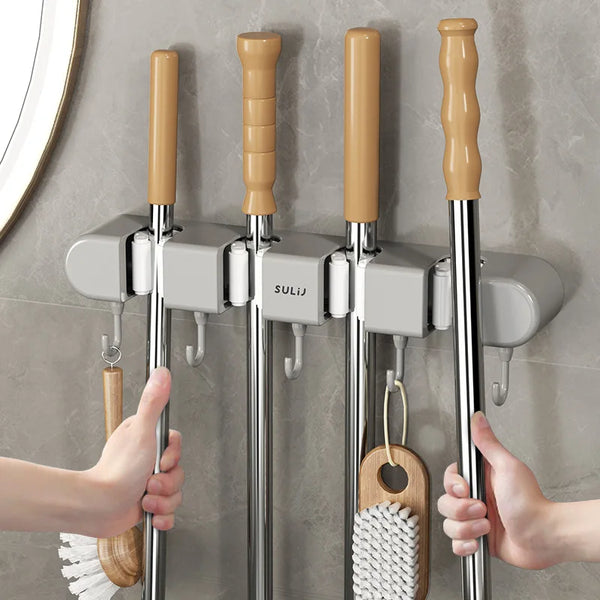 Hands are installing a wall-mounted mop and broom holder with an adhesive mount onto a gray tiled wall.