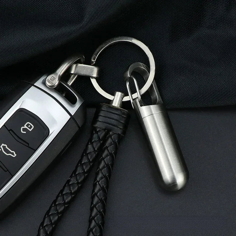 A stainless steel waterproof pill capsule attached to a keychain with a black leather cord.