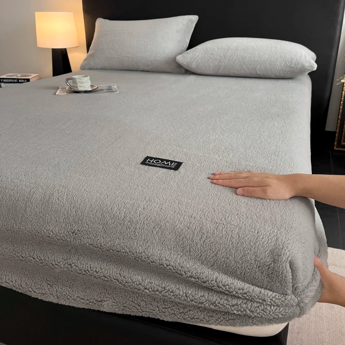 A gray plush mattress cover is shown on a bed, with a hand touching the fabric.