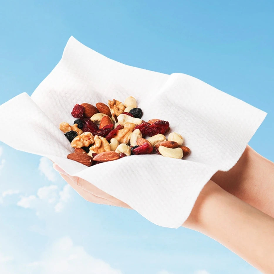 Una mano sostiene una toalla facial desechable blanca llena de frutos secos y fruta seca contra un cielo azul.