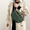 A woman wearing a beige trench coat holds a dark green woven pattern waist bag with a gold chain.