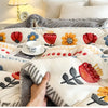 A hand is touching a white and gray plush throw blanket featuring floral embroidery on a bed.