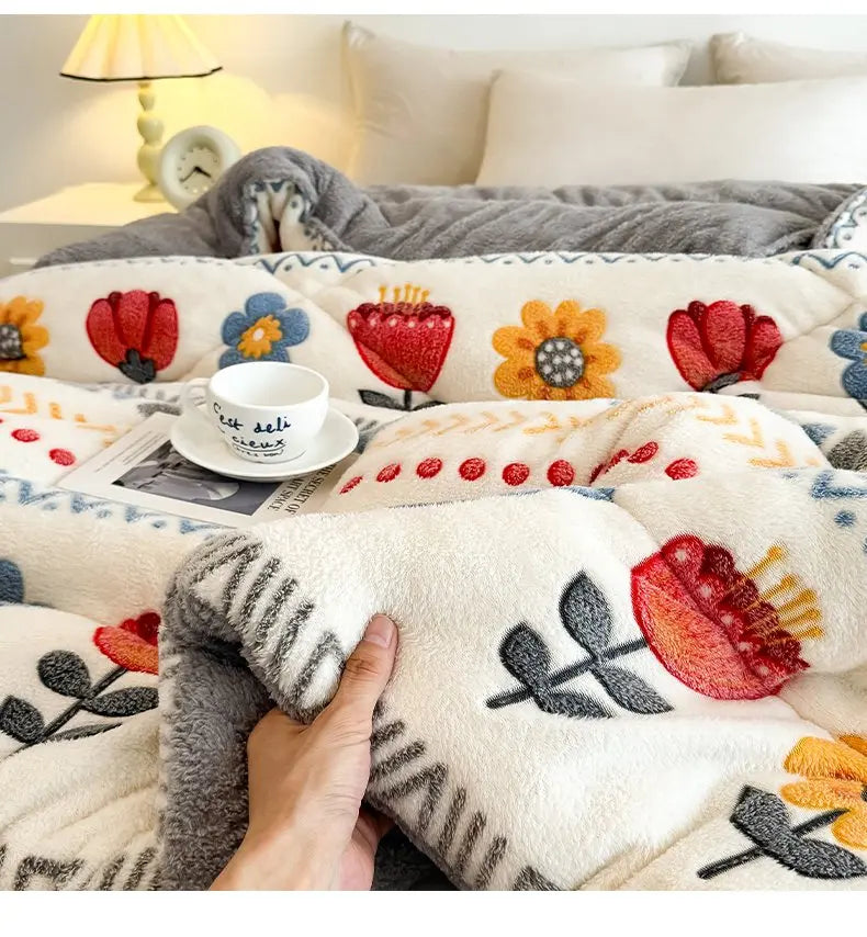 A hand is touching a white and gray plush throw blanket featuring floral embroidery on a bed.