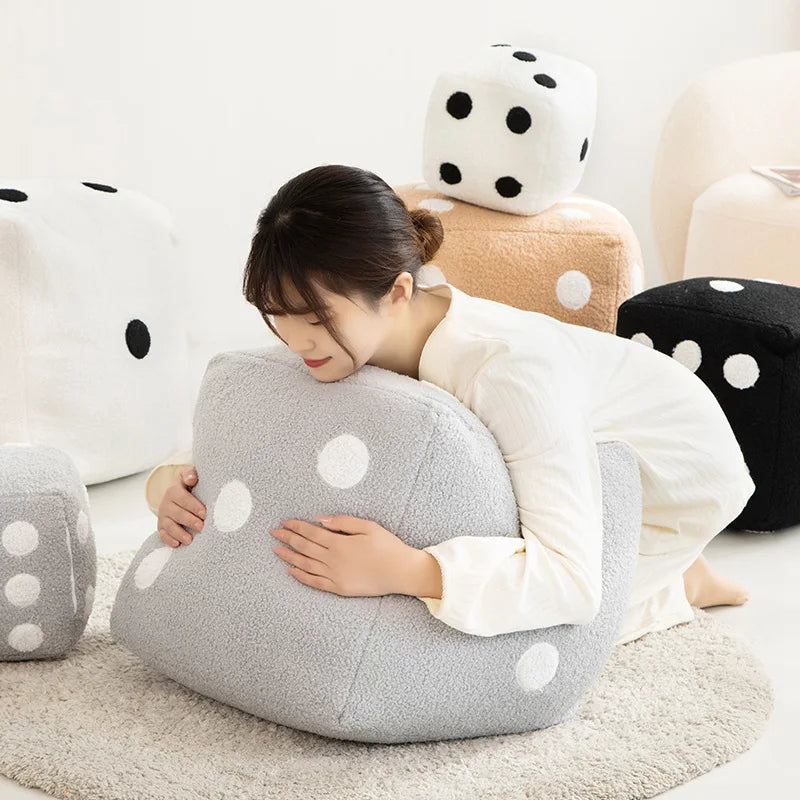 A woman rests her head on a large gray plush pillow decorated with white polka dots, surrounded by other soft, geometric cush