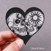 A black heart-shaped patch embroidered with two detailed skulls, shown being held by a hand.
