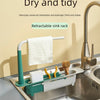 A white and green retractable sink rack holds a wet towel and sponge in a stainless steel kitchen sink.