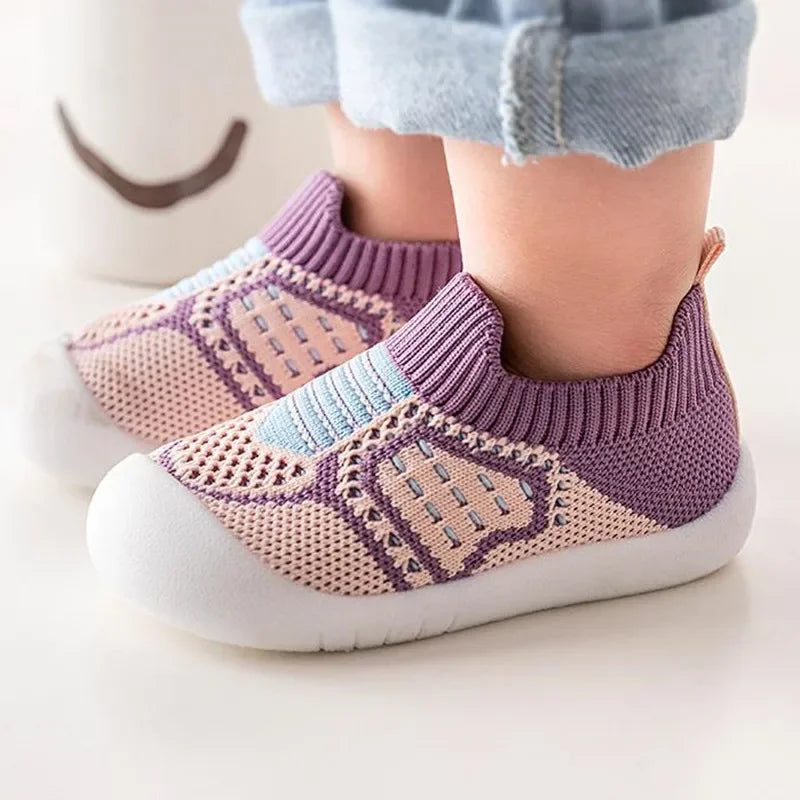 A pair of purple and beige knitted baby shoes with a soft sole resting on a white surface.