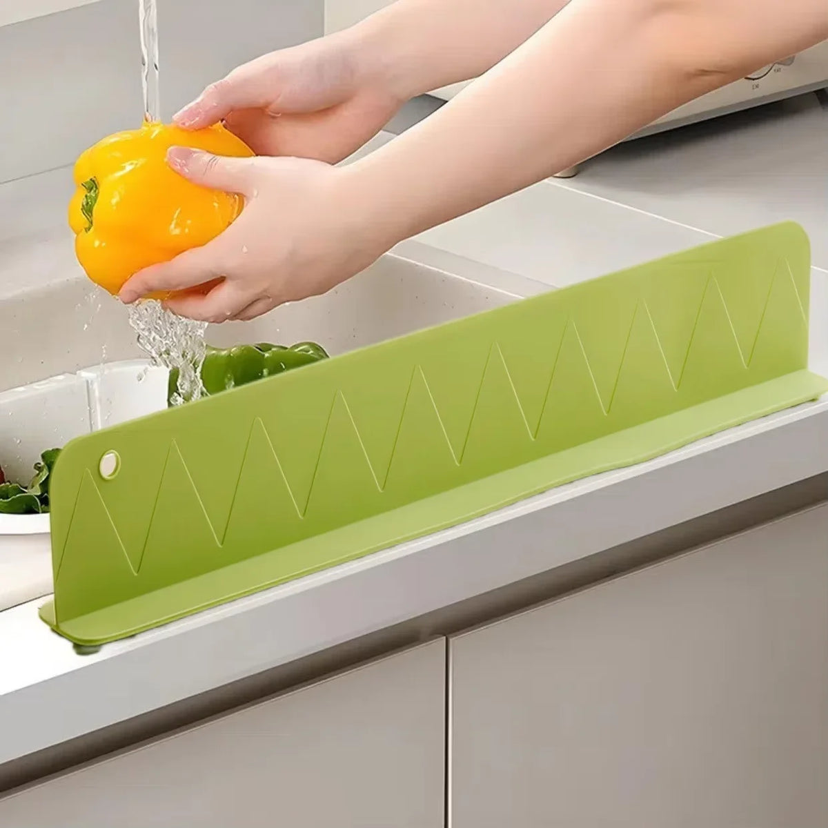 A hand is rinsing a yellow bell pepper under a flexible silicone sink splash guard in a kitchen sink.