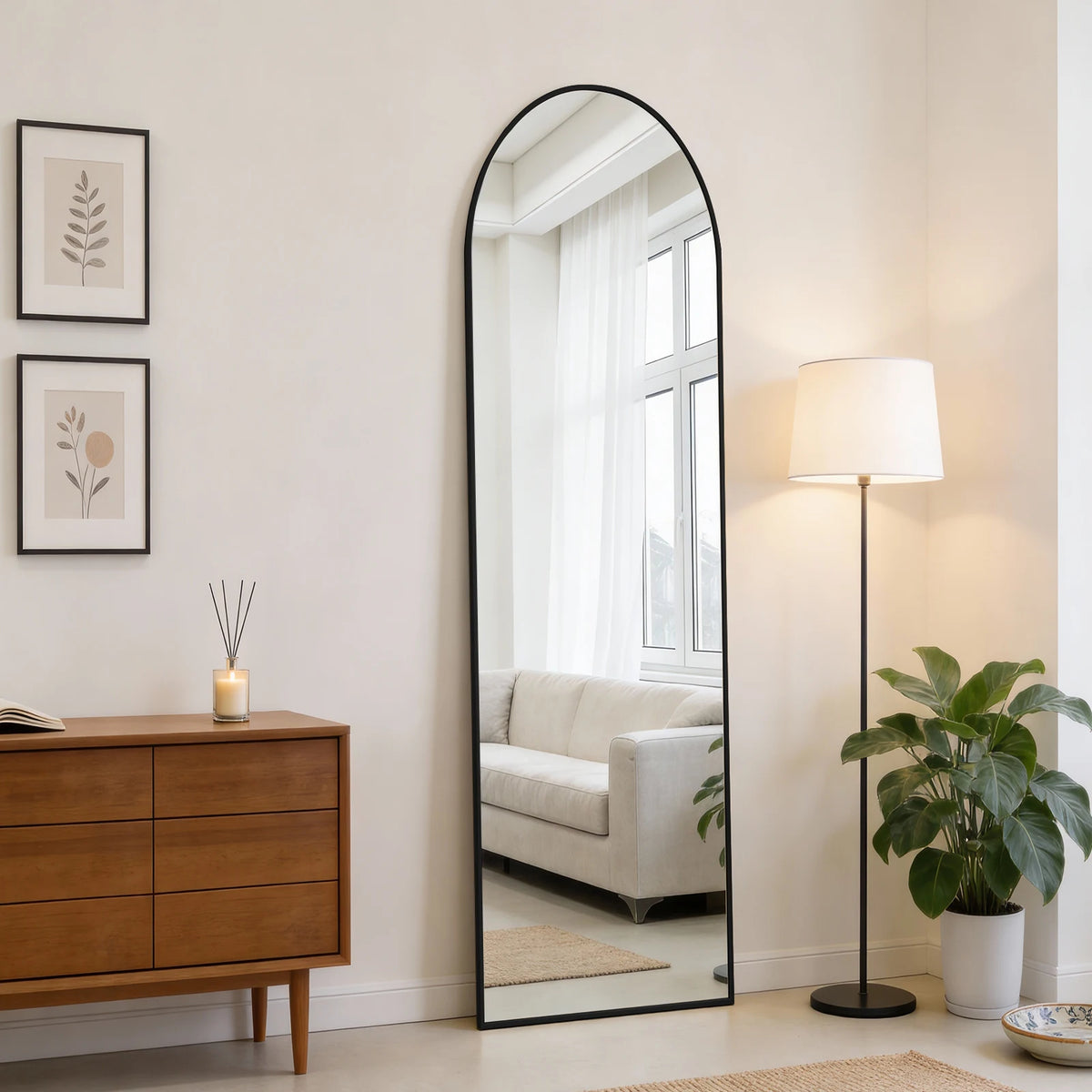 A full length oval wall mirror with a black frame reflects a bright room and a window.