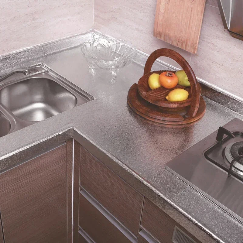 A kitchen countertop with a stainless steel sink, wooden bowl of fruit, and a stovetop area.