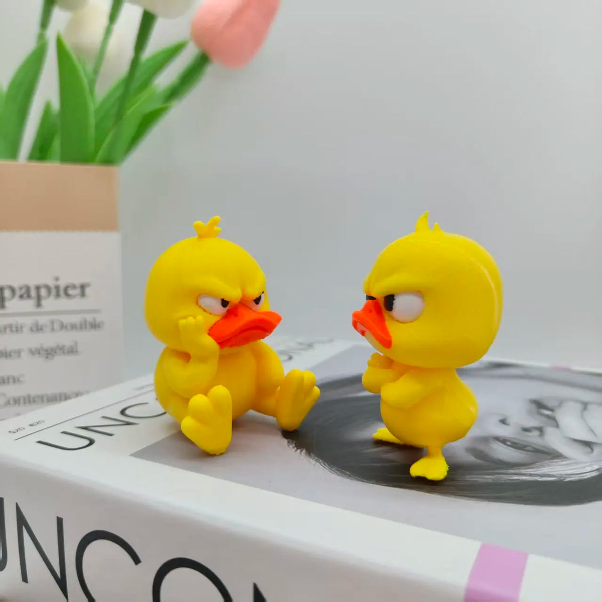 A yellow 3D printed duck statue decoration sitting on a white box next to a pink tulip.