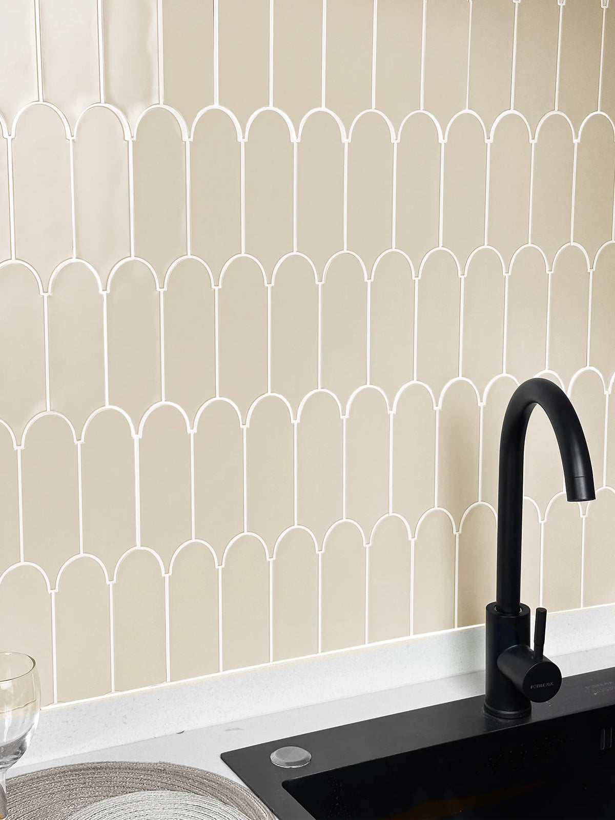 A kitchen backsplash featuring white arched tiles on a beige wall above a black countertop and faucet.
