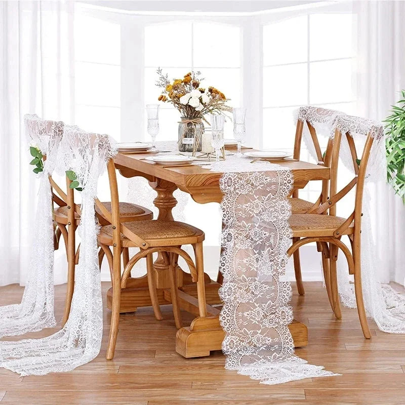 A wooden dining table set for a meal with embroidered floral lace table runners and sheer white fabric on a bright windowsill