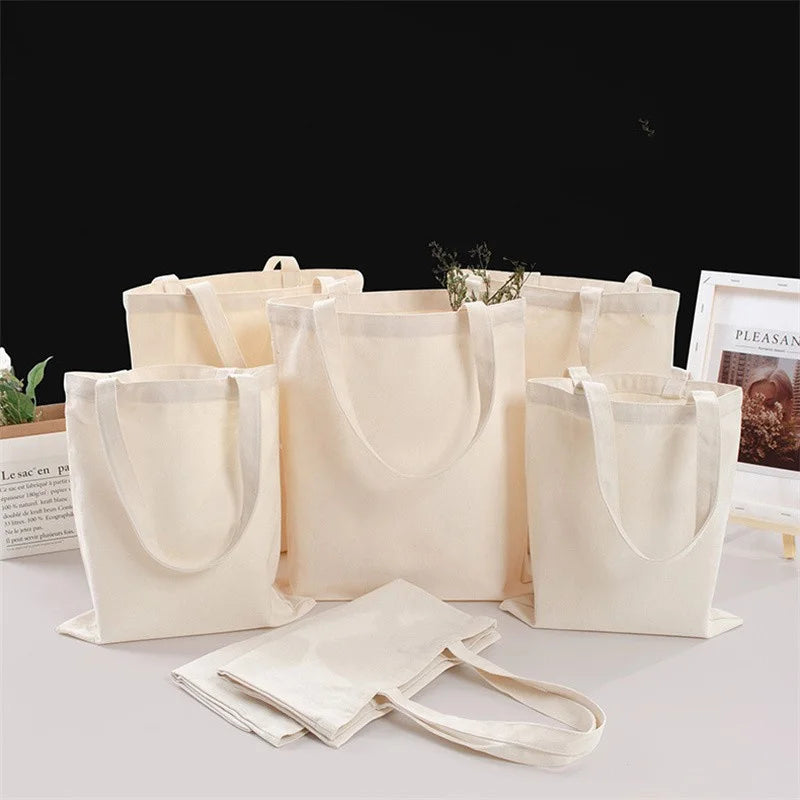 Several natural canvas shopping tote bags in beige and off-white colors are displayed on a white surface.