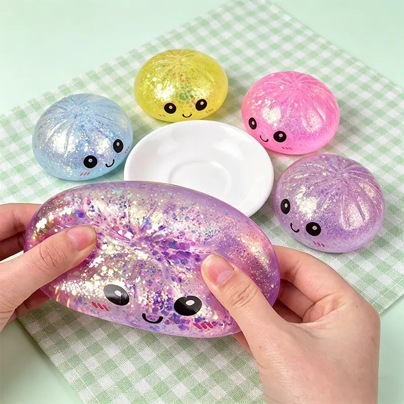 Hands hold a purple glitter dumpling squeeze toy next to several other colorful glitter toys on a green checkered cloth.