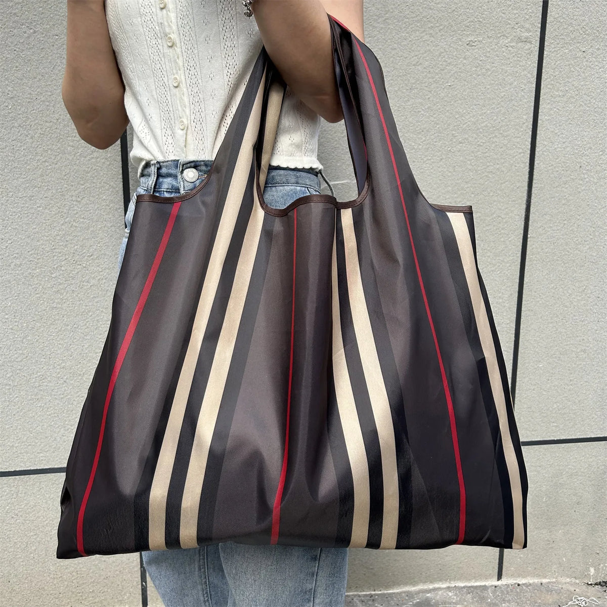 A person holds a dark brown and beige striped foldable oxford cloth tote bag against a gray wall.
