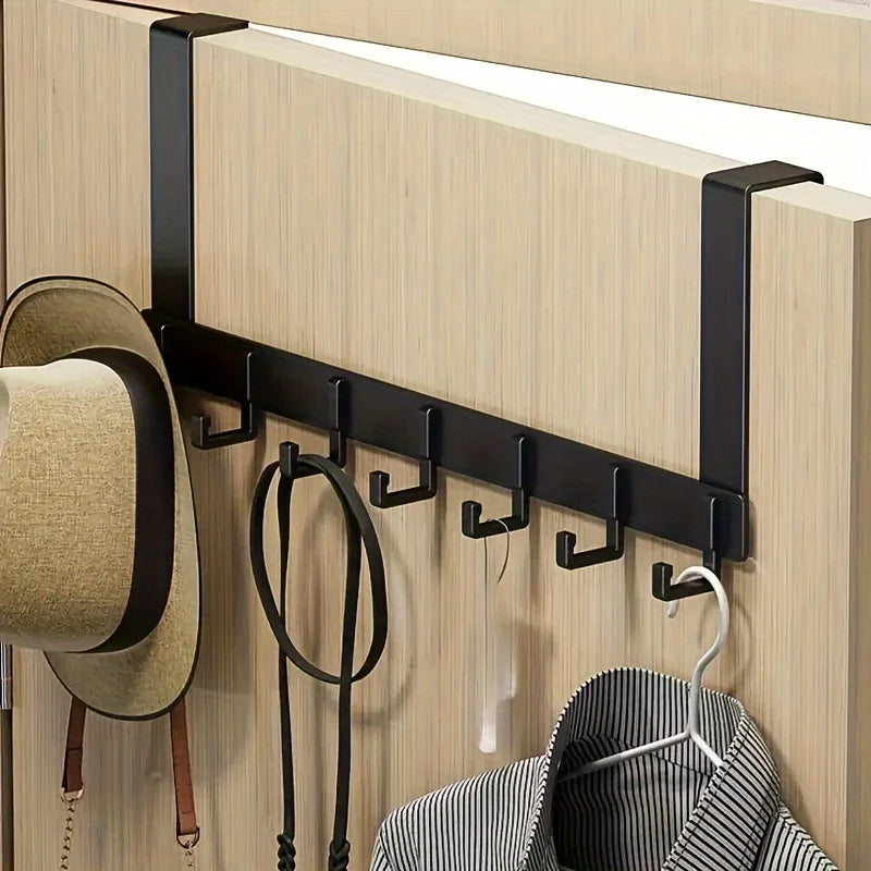 A black over-the-door coat rack with six hooks mounted on a light wood panel, holding a hat and a striped garment.