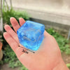 A hand holds a clear blue ice cube shaped stress relief toy with a snowflake design against a blurred green outdoor backgroun