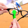 A hand is using a green brush to clean the lens of black eyeglasses.