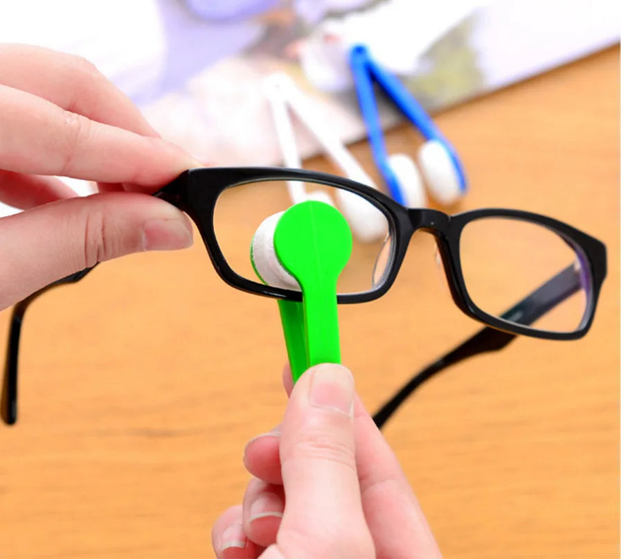 A hand is using a green brush to clean the lens of black eyeglasses.