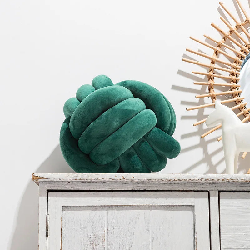 A teal green knitted velvet throw pillow resting on a white wooden shelf next to a light-colored decorative sunburst object.