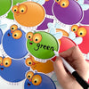 A hand writes the word green on a dry erase fly swat game card featuring an orange-colored character.