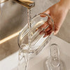 Eine Hand gießt klare Flüssigkeit aus einem goldenen Wasserhahn in eine elegante Glasseifehalter auf einer weißen Arbeitsplat