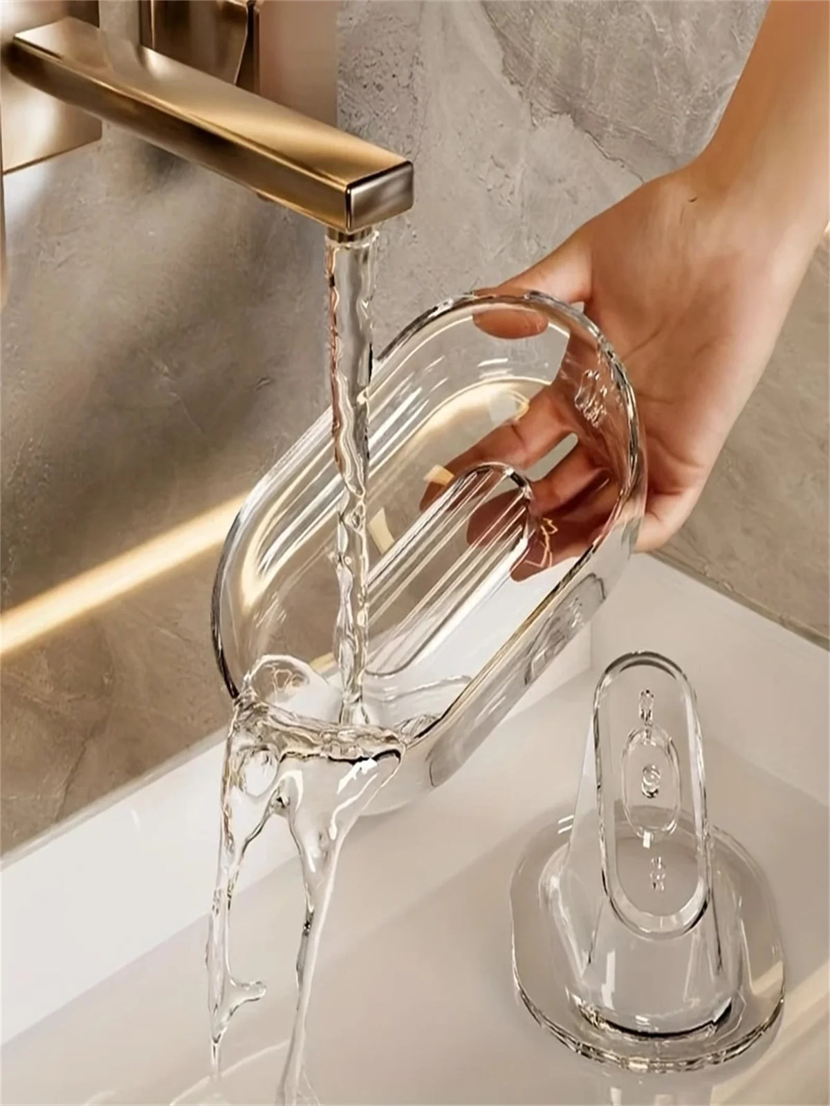 Eine Hand gießt klare Flüssigkeit aus einem goldenen Wasserhahn in eine elegante Glasseifehalter auf einer weißen Arbeitsplat