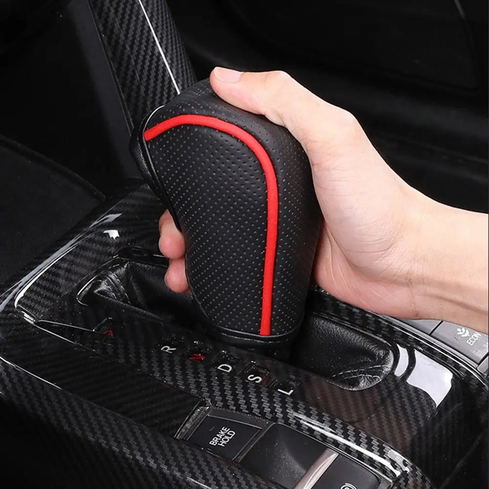 A hand is holding a black gear shift knob cover with a red accent, shown in the context of a vehicle interior.