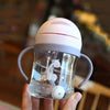 A hand holds a clear plastic straw cup with handles and a white lid, featuring a small bear design.