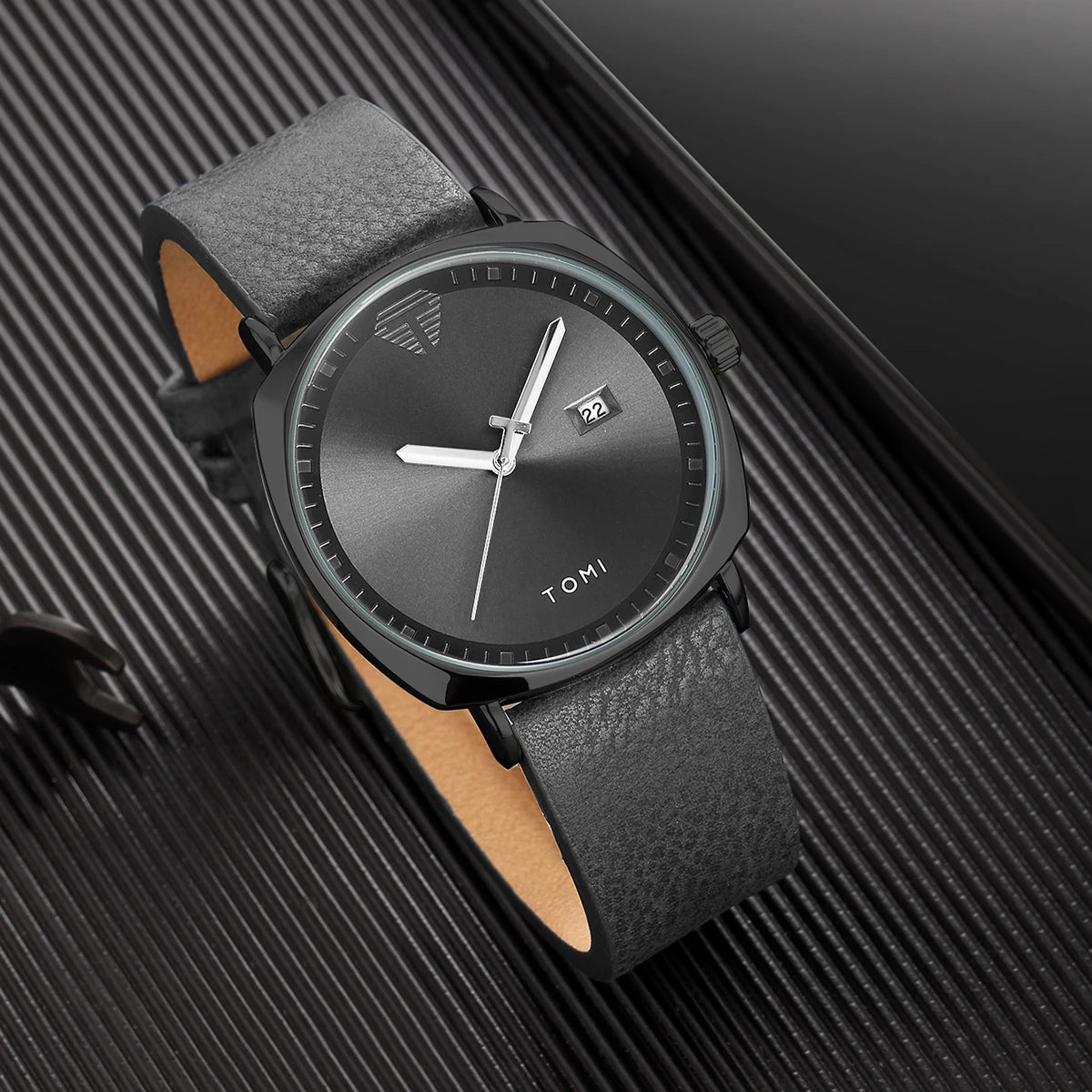 A black watch with a silver dial and brown leather strap is displayed on a dark surface.