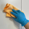 A hand wearing a blue nitrile glove is holding an orange woven cloth against a white surface.